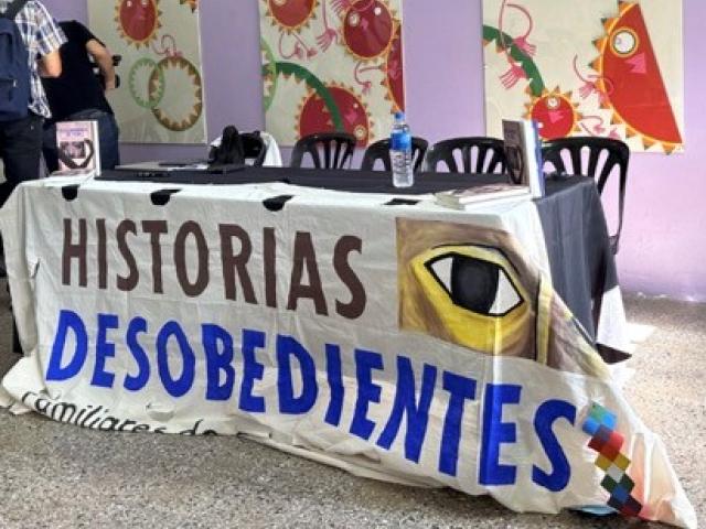 Sign reading "HISTORIAS DESOBEDIENTES" is draped over a table with five chairs..